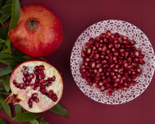 top view of pomegranate berries in bowl with pomegranate half and whole one with leaves on bordo background